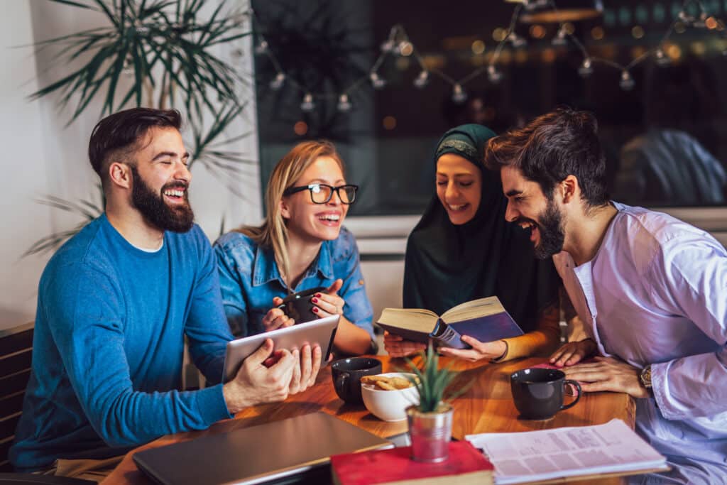 Three people sit a table in a cafe. They are mixed race, laughing and talking. Showing social and cultural sensitivity in a safe workplace.