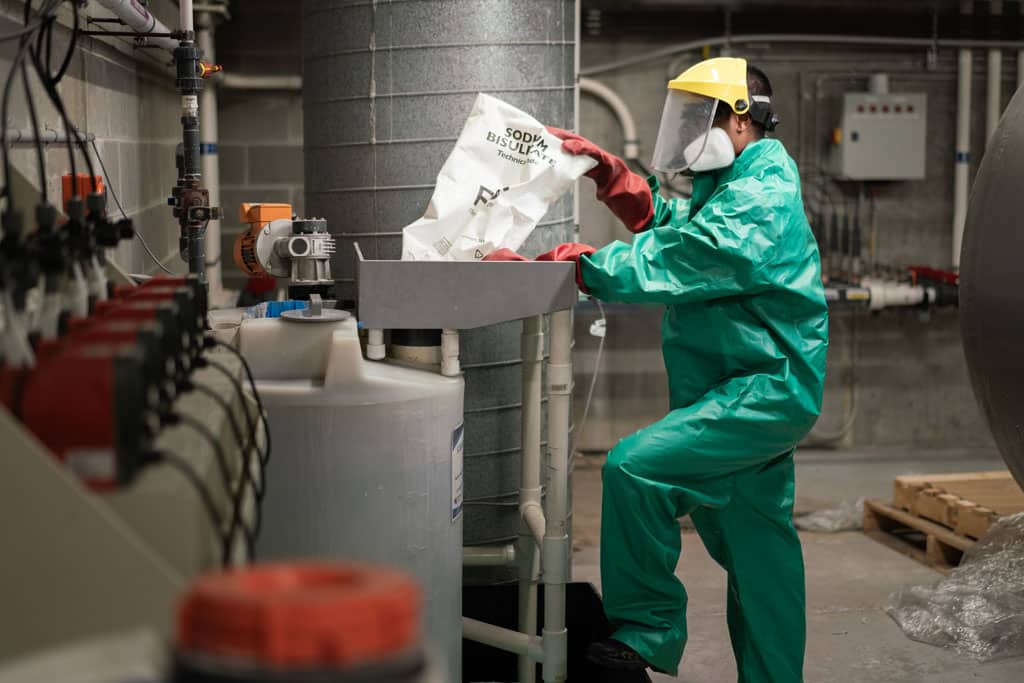 a man in a green suit and face mask putting a bag of chemicals in a pump for a swimming pool