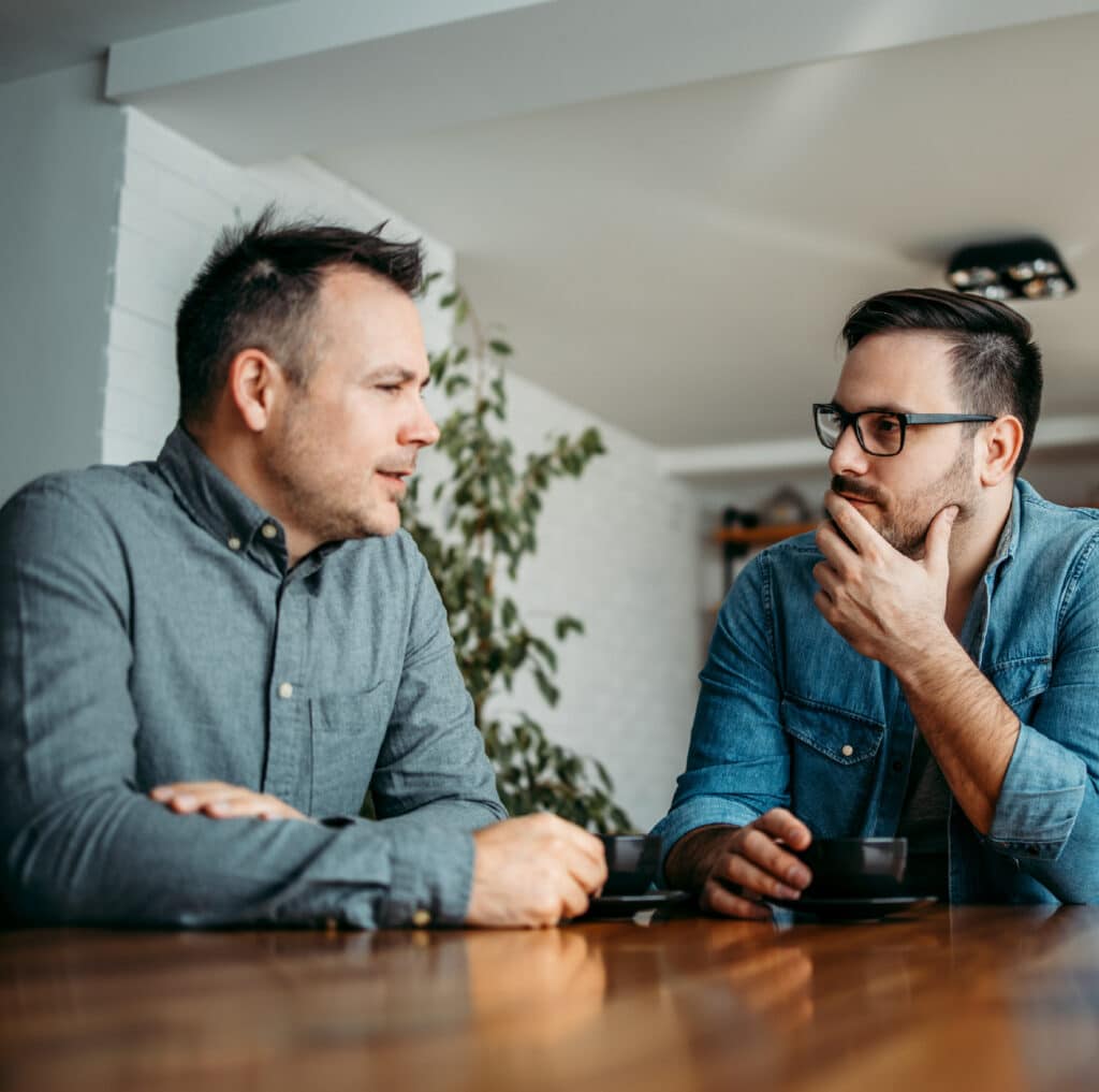 Two men sit at a table and talk seriously. They manage conflict.