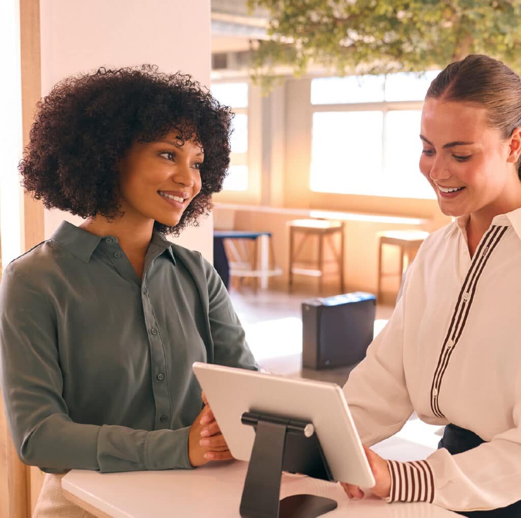 A lady serves a female customer and works to enhance customer service experiences. They stand at the reception desk in a foyer.